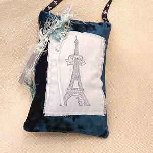Könnte beinhalten: Eine kleine blaue Samt-Tasche mit einem weißen Stoff-Patch, auf dem eine Schwarzweiß-Illustration des Eiffelturms mit dem Text "Paris" und "La Tour Eiffel" abgebildet ist. Die Tasche hat ein schwarzes Band mit Fleur-de-lis-Akzenten.