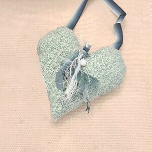 Puede incluir: Un adorno de tela verde en forma de corazón con un lazo de cinta azul y un adorno de plumas blancas y azules.