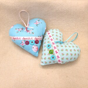 May include: Two blue fabric heart-shaped ornaments with pink and white floral patterns. One heart has a white ribbon with the text "cherish" repeated three times. Both hearts have buttons.
