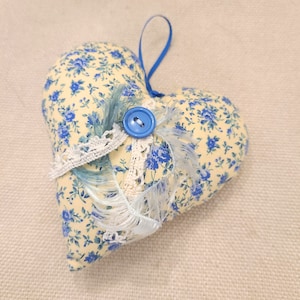 Puede incluir: Una almohada en forma de corazón amarilla con un estampado floral azul y un botón azul. La almohada tiene una cinta azul y un borde de encaje blanco.