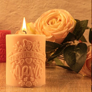Puede incluir: Una vela de pilar beige con el texto "World's Best Mama" en relieve en el lateral. La vela está encendida y rodeada de rosas artificiales.