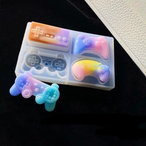 May include: A clear silicone mould with four different shapes of video game controllers. The mould is filled with colourful resin, creating a rainbow effect.