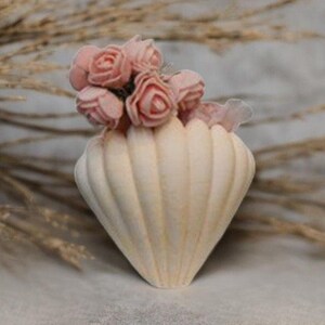 May include: A small, off-white, seashell-shaped vase with vertical ridges, holding a cluster of small, pink roses. The vase is set against a soft, neutral background, creating a delicate and natural aesthetic. The arrangement evokes a sense of floral elegance.