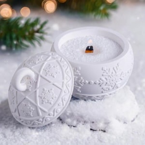 Op de afbeelding: Een witte, ronde kaars met een deksel, versierd met sneeuwvlokpatronen. De kaars is aangestoken, met een houten lont. Het deksel ligt opzij. De kaars staat op een besneeuwd oppervlak, met een vage achtergrond van kerstverlichting en groen.