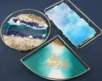 May include: Three resin trays with gold metal frames. The trays are filled with blue, green, and white resin. The round tray has a beach scene with sand, shells, and blue glitter. The square tray has a blue and white swirl design. The triangular tray has a green and white swirl design with the word "Lucky" written in gold.
