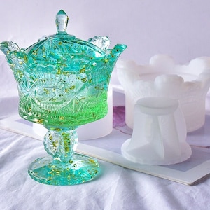 Puede incluir: Un plato de dulces de vidrio verde con un diseño de hoja de oro y una tapa. El plato está sobre una base de pedestal blanco. El plato está al lado de un molde de silicona blanco.