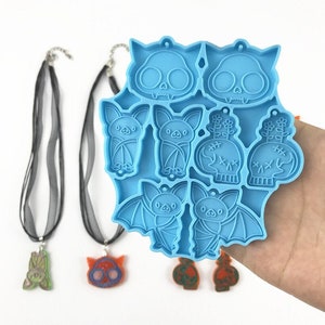 Puede incluir: Un molde de silicona azul con ocho formas diferentes de tem&aacute;tica de Halloween, incluyendo murci&eacute;lagos, calaveras y una planta. El molde est&aacute; en una mano, y hay varias piezas de resina terminadas al lado.