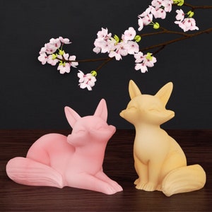 Peut inclure: Deux figurines de renard en céramique, l'une rose et l'autre jaune, avec les yeux fermés et une expression souriante. Les figurines sont assises sur une surface en bois avec une branche de fleurs de cerisier roses en arrière-plan.