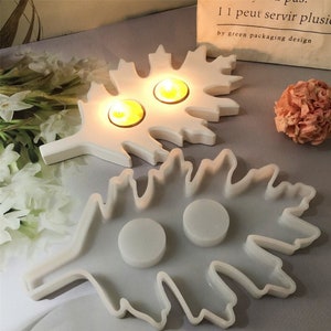Op de afbeelding: Twee witte, bladvormige kaarsenhouders. De bovenste houder heeft twee kleine kaarsverzonkingen en de onderste houder heeft twee grotere verzonkingen. De houders zijn gemaakt van een glad, wit materiaal.