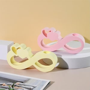 Op de afbeelding: Twee decoratieve objecten in de vorm van een oneindigheidssymbool, een geel en een roze, met een hartvormige uitsparing aan de bovenkant. De objecten zijn gemaakt van hout en hebben een gladde afwerking.