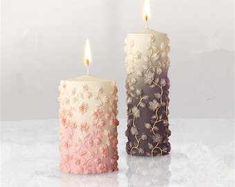 Könnte beinhalten: Zwei dekorative Kerzen mit floralen Mustern. Die Kerze links ist rosa und weiß, die Kerze rechts ist grau und weiß.