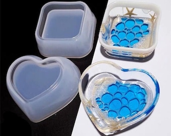 Puede incluir: Tres moldes de resina transparente para manualidades. Un molde cuadrado, un molde en forma de corazón y un molde en forma de corazón con un diseño azul y blanco.