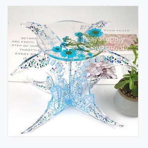 May include: A clear resin stand with a geometric design and a round top. The stand has four legs that are shaped like crescent moons. The top of the stand is decorated with blue flowers and glitter. The stand is sitting on a book with the text "WIND AGAIN" visible.