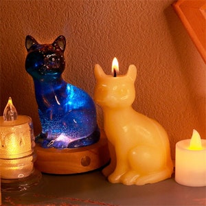 May include: Two cat-shaped candles, one blue glass with glitter and a lit LED, the other a solid yellow candle with a lit wick. Both are sitting on a wooden base.