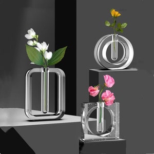 Könnte beinhalten: Drei transparente Glasvasen in verschiedenen Formen, jede mit einem einzelnen Stiel Blumen. Die Vasen stehen auf einer schwarzen Oberfläche. Die Blumen sind weiß, gelb und rosa.