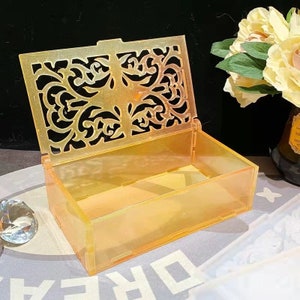 Op de afbeelding: Een doorschijnende gele decoratieve doos met een gesneden bloemenpatroon op de deksel. De doos is open en het interieur is zichtbaar.