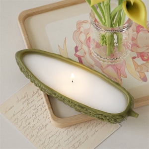 Puede incluir: Una vela verde con forma de pepino con un interior de cera blanca y una mecha encendida. La vela está sobre una bandeja de madera con un diseño floral.
