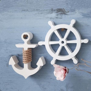 May include: White wooden nautical decor set with a ship's wheel and an anchor with rope detail. The decor is on a blue wooden background.