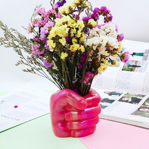 May include: A pink iridescent hand-shaped vase with a bouquet of dried flowers in shades of purple, yellow, and white.