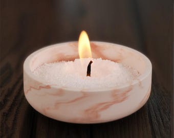 Puede incluir: Una vela blanca con un soporte de mármol rosa. La vela está encendida y la mecha es visible.