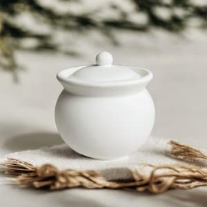Pode incluir: Um pequeno pote de cerâmica branco com tampa e corpo arredondado. A tampa tem um pequeno botão redondo no topo. O pote está sobre um pano branco com um elemento decorativo castanho tecido.