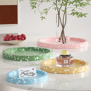 May include: Four round trays in different colors, pink, green, yellow, and blue, with white splatter paint. The trays are on a white surface. The word "Britney" is written on a white card on the blue tray.