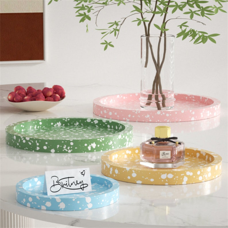 May include: Four round trays in different colors, pink, green, yellow, and blue, with white splatter paint. The trays are on a white surface. The word "Britney" is written on a white card on the blue tray.