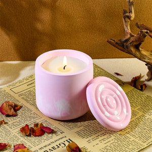 May include: A pink candle with a white lid. The candle is in a cylindrical container with a white design. The lid has a white swirl design.