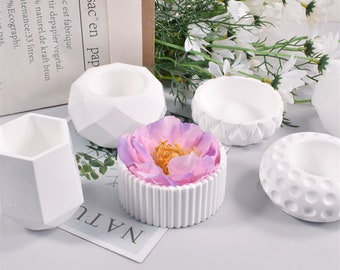May include: Five white geometric concrete candle holders of different shapes and sizes. The holders are arranged on a white surface with a pink flower in the center.