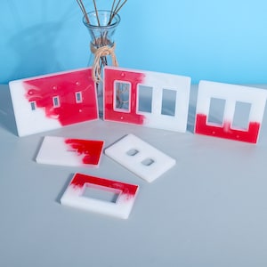 May include: Set of six white and red resin light switch covers. The covers have a marbled effect with red and white swirls. The covers are in various sizes and shapes, including single, double, and triple switch covers.