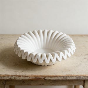 May include: A white ceramic bowl with a ruffled, scalloped edge. The bowl is round and sits on a light-colored wooden table. The bowl's design features a series of vertical, curved ridges that create a textured surface. The bowl is empty.