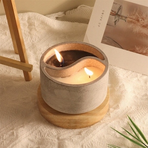 May include: A gray concrete candle holder with a yin yang design. The candle holder has two compartments, one with a black candle and one with a white candle. Both candles are lit.
