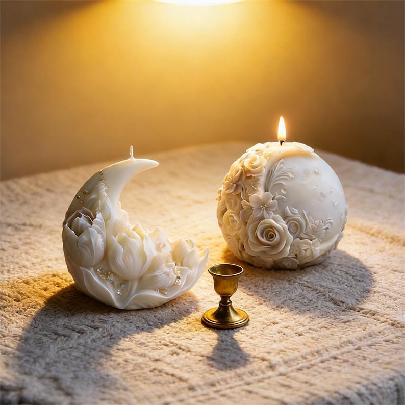 May include: Two decorative candles, one crescent moon-shaped and the other spherical, both in ivory with floral designs. A small, gold-colored candle holder sits between them. The spherical candle is lit, casting a warm glow.