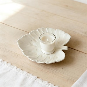 May include: A white candle in a clear glass votive sits on a white, leaf-shaped ceramic dish. The dish has detailed veining and a small handle. The candle is lit, and the background is a light-colored wooden surface.