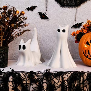 May include: Two white cat-shaped ghost figurines with black eyes, set on a table decorated for Halloween. A carved orange pumpkin with a lit face sits to the right. Black bat cutouts and floral arrangements complete the spooky scene.