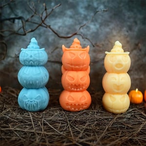Peut inclure: Trois décorations empilées en forme de citrouille, bleues, oranges et jaunes. Chaque citrouille a un visage sculpté et est surmontée d'un chapeau de sorcière. Les décorations sont sur un lit de paille, avec un fond sombre.