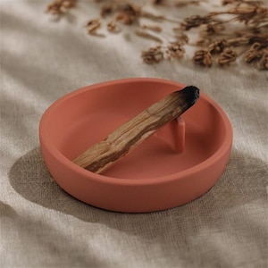 以下が含まれることがあります： テラコッタ色のセラミック香立てと燃えたパロサントの棒。香立ては丸く、お香を支える小さな隆起したサポートがあります。パロサントの棒は茶色で濃い縞模様があり、端は黒くなっています。