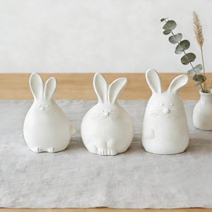 May include: Three white ceramic bunny ornaments with detailed features, including long ears and whimsical expressions. The bunnies are arranged on a grey linen table runner, creating a charming Easter or spring-themed display.