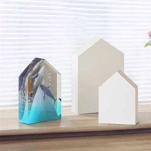 Op de afbeelding: Een transparant harsbeeld van walvissen die in een blauwe oceaan zwemmen. Het beeld is gevormd als een huis en wordt getoond op een houten oppervlak met twee witte, huisvormige houten objecten.