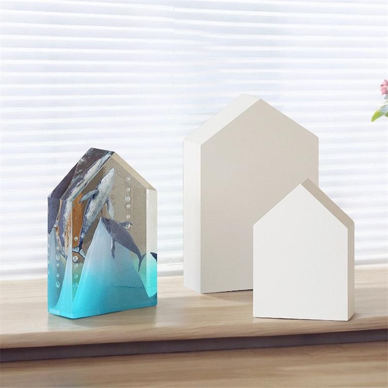 Op de afbeelding: Een transparant harsbeeld van walvissen die in een blauwe oceaan zwemmen. Het beeld is gevormd als een huis en wordt getoond op een houten oppervlak met twee witte, huisvormige houten objecten.