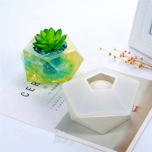 May include: A white silicone mold for making a geometric planter. The mold is shaped like a pentagon with a hexagonal opening in the center. A finished planter made with the mold is shown in the background, filled with a succulent plant.