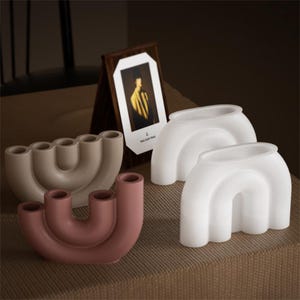 May include: A collection of modern ceramic vases in various colors. The vases have a unique arched design, with one in a muted pink, one in beige, and two in white. A framed photograph is in the background.