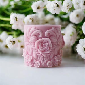 May include: A cylindrical, light pink candle with a detailed floral design. The candle features a central rose surrounded by ornate leaves and smaller roses. White flowers are in the background.