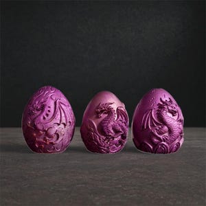 Pode incluir: Três ovos de dragão roxos com detalhes intrincados esculpidos. Cada ovo apresenta um design de dragão diferente, com asas e escamas. Os ovos são exibidos em uma superfície escura, criando um tema de fantasia.