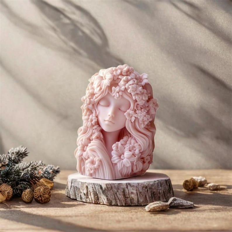 May include: A pink sculpture of a woman's face with closed eyes and flowing hair adorned with flowers. The sculpture is mounted on a small, round wooden base. The overall aesthetic is soft and feminine.