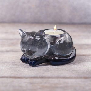 Può includere: Un portacandele trasparente a forma di gatto con una candela accesa all'interno. Il gatto è in posizione di riposo, con le zampe ripiegate. Il portacandele è realizzato in un materiale trasparente e scintillante con dettagli blu scuro.