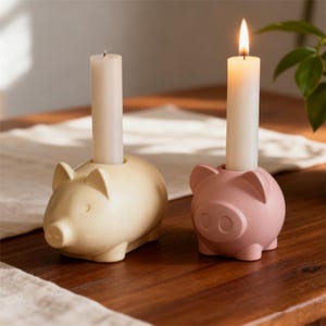 Puede incluir: Dos candelabros de cerámica con forma de cerdo, uno crema y otro rosa, cada uno con una vela blanca. La vela del soporte rosa está encendida. Los candelabros están sobre una superficie de madera.