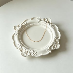 Pode incluir: Um delicado colar de corrente dourada repousa sobre um prato decorativo branco e ornamentado. O prato tem uma borda ondulada e enfeites florais. O colar é simples e elegante.