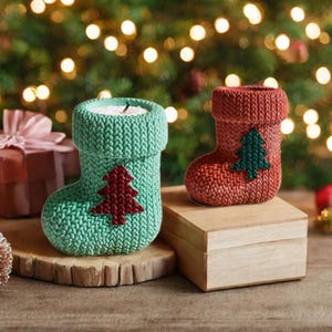 May include: Two knitted Christmas stocking candle holders. One is teal with a red tree, the other is red with a green tree. They are on wooden surfaces, with a blurred background of Christmas lights.