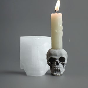 May include: A lit white candle in a gray skull-shaped candle holder. A white, skull-shaped mold is to the left. The candle holder has detailed facial features. The candle is burning with a yellow flame. The background is a solid gray.